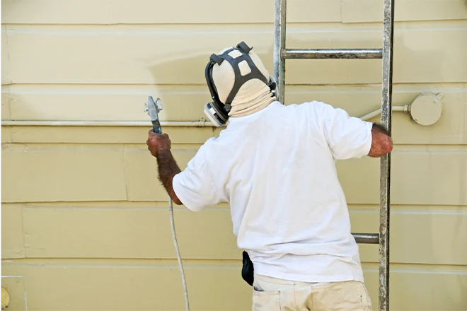A man on a ladder uses paint spray gun on house siding